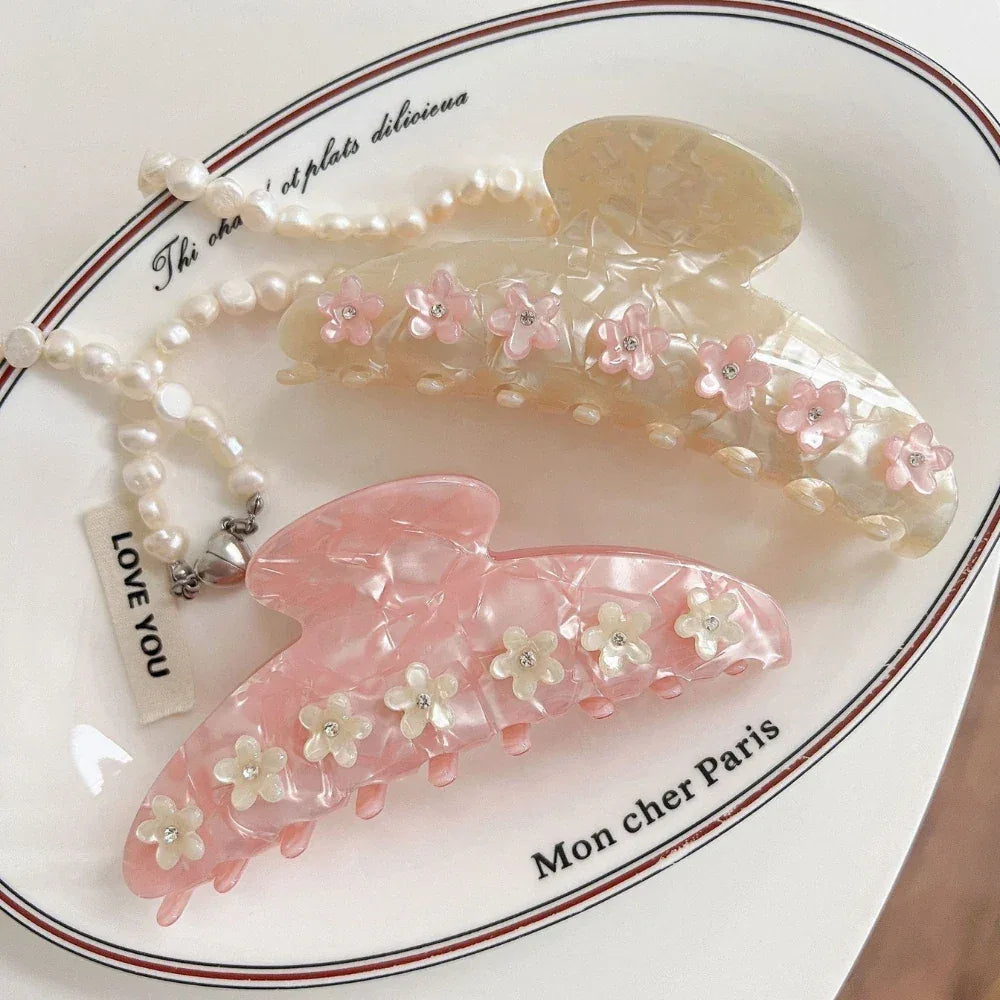 Two decorative claw clips, one pink and one cream, adorned with floral appliques and gemstones, placed on a decorative dish with a pearl necklace.