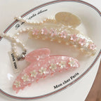 Two decorative claw clips, one pink and one cream, adorned with floral appliques and gemstones, placed on a decorative dish with a pearl necklace.