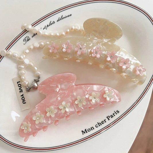 Two decorative claw clips, one pink and one cream, adorned with floral appliques and gemstones, placed on a decorative dish with a pearl necklace.