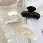 Three decorative claw clips arranged on a white textured surface: one cream-colored with purple floral appliques and gold beads, one black with black floral and butterfly appliques and rhinestones, and one iridescent white with white floral appliques and rhinestones.