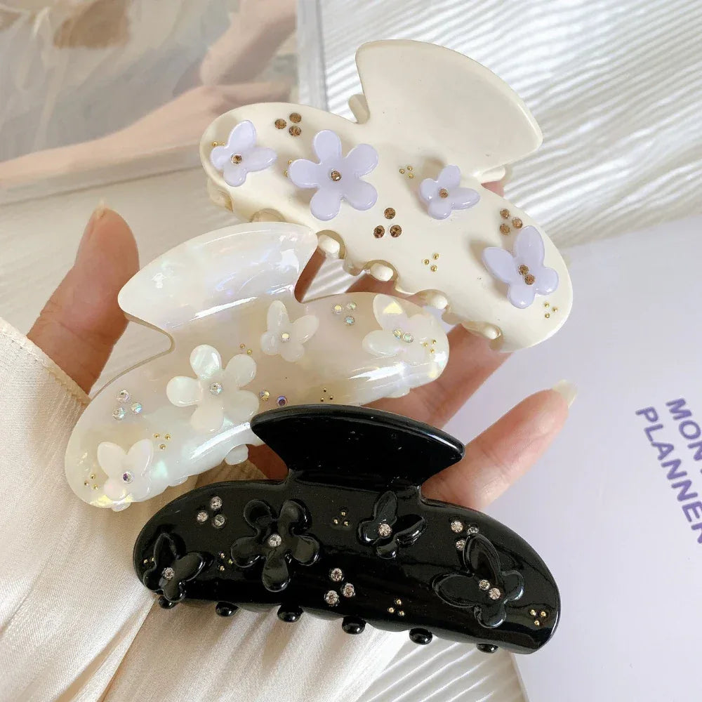 A hand holding three decorative claw clips: a black clip with floral and butterfly appliques and rhinestones, a cream clip with purple floral appliques and gold beads, and an iridescent white clip with white floral appliques and rhinestones.