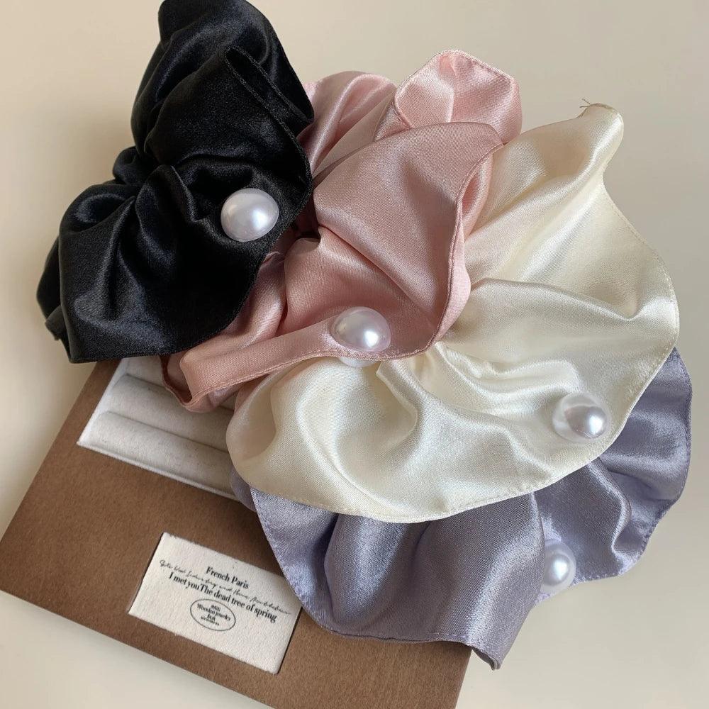 A collection of four satin scrunchies in black, pink, cream, and gray, each with a pearl accent, arranged on a brown box.