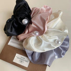 A collection of four satin scrunchies in black, pink, cream, and gray, each with a pearl accent, arranged on a brown box.