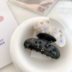 A black claw clip with black floral and butterfly appliques and clear rhinestones, placed inside a white ceramic bowl next to a cream-colored claw clip with purple floral appliques and gold beads.
