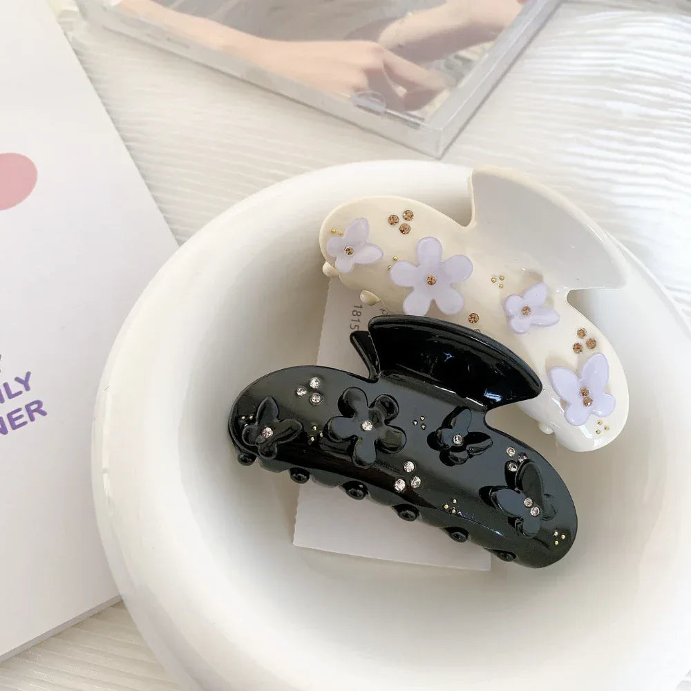A black claw clip with black floral and butterfly appliques and clear rhinestones, placed inside a white ceramic bowl next to a cream-colored claw clip with purple floral appliques and gold beads.