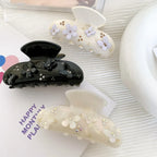 Three decorative claw clips displayed on a white surface: a black clip with floral and butterfly appliques and rhinestones, a cream clip with purple floral appliques and gold beads, and an iridescent white clip with white floral appliques and rhinestones.