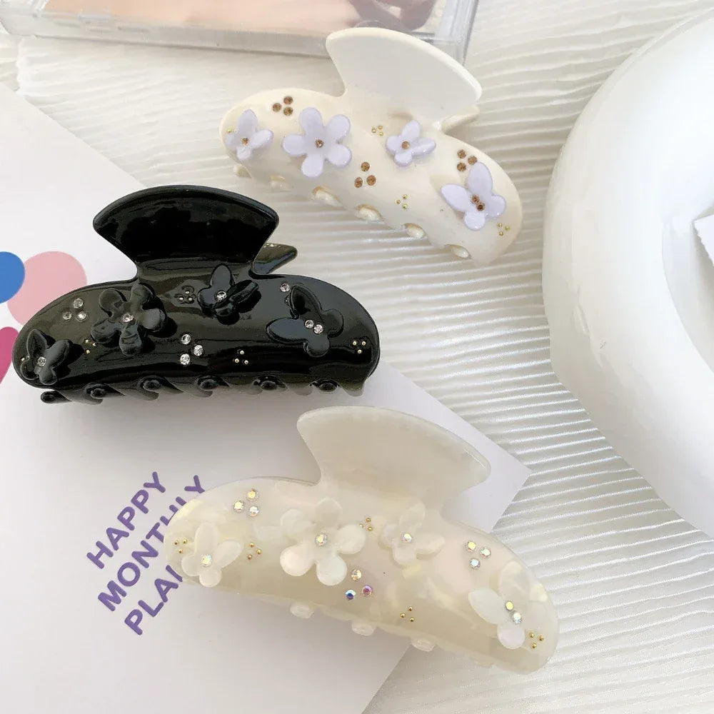 Three decorative claw clips displayed on a white surface: a black clip with floral and butterfly appliques and rhinestones, a cream clip with purple floral appliques and gold beads, and an iridescent white clip with white floral appliques and rhinestones.