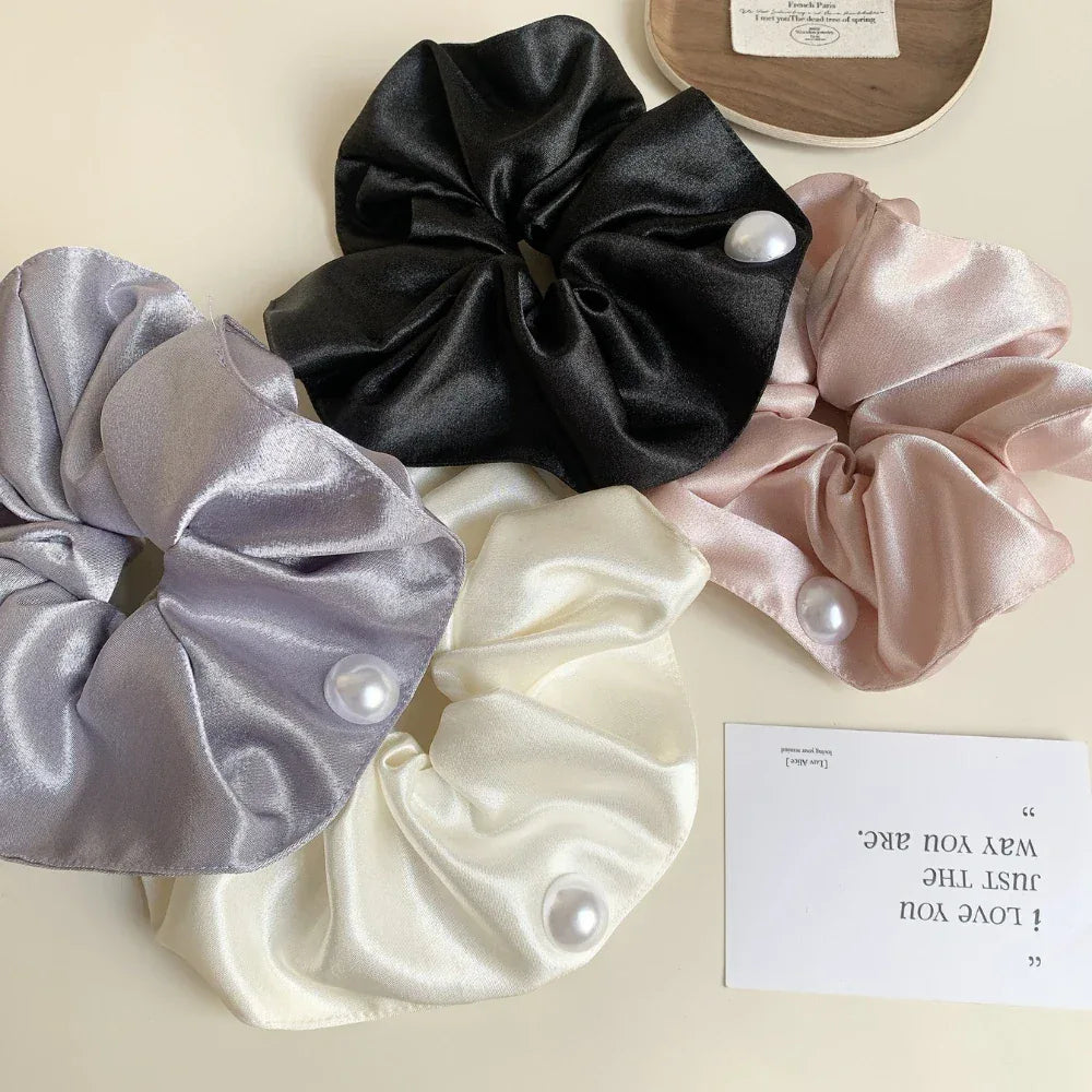Four satin scrunchies in black, pink, cream, and gray, each with a pearl accent, laid out on a light surface with a small card.