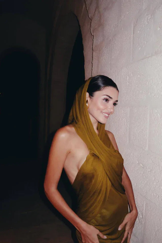 Woman smiling and wearing a draped olive green halter dress with a hood, outdoors at night.