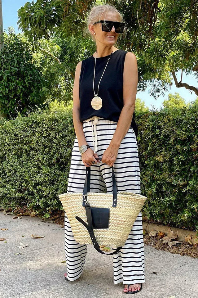 Woman wearing a black sleeveless top and black and white striped wide leg pants, holding a straw tote bag.