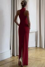 Back view of a woman wearing a burgundy sleeveless halter maxi dress with a gold-tone hardware detail and a back slit, standing in a room with curtains.