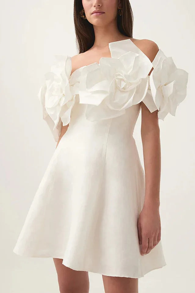 Model wearing a white off-the-shoulder mini dress with large floral embellishments on the shoulders and white heels.