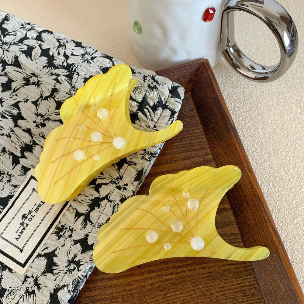 Two yellow ginkgo leaf-shaped hair claw clips resting on a black and white floral fabric and a wooden tray.