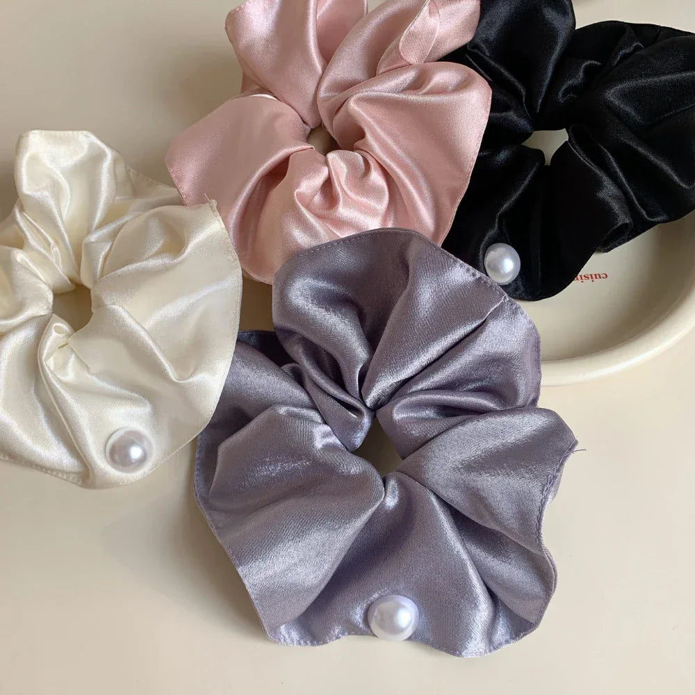 A top-down view of five satin scrunchies in black, pink, cream, and gray, each with a pearl accent, on a light surface.