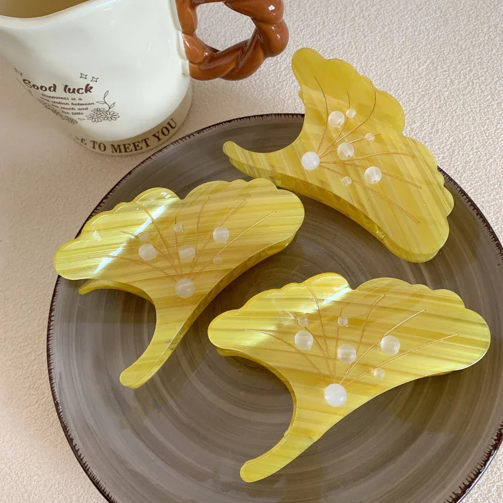 Three yellow ginkgo leaf-shaped hair claw clips arranged on a ceramic plate, with a mug in the background.
