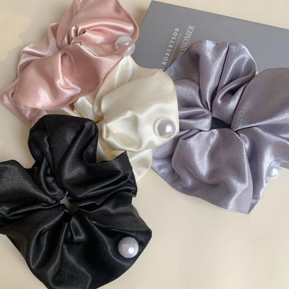Four satin scrunchies in black, pink, cream, and gray, each with a pearl accent, arranged on a light surface with a book.