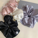 Four satin scrunchies in black, pink, cream, and gray, each with a pearl accent, arranged on a light surface with a book.
