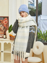 Woman wearing a grey knit sweater with blue and white geometric pattern, a light blue beanie, and a chunky striped scarf, looking at the camera.