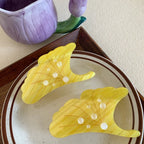 Two yellow ginkgo leaf-shaped hair claw clips on a plate, with a purple floral mug in the background.