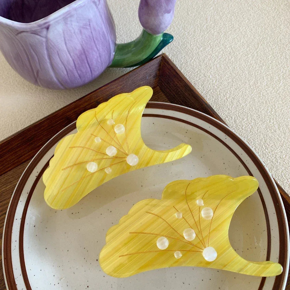 Two yellow ginkgo leaf-shaped hair claw clips on a plate, with a purple floral mug in the background.