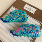 A pair of peacock hair claw clips, one with pink accents on the feathers and the other with orange accents, nestled in a jewelry box.