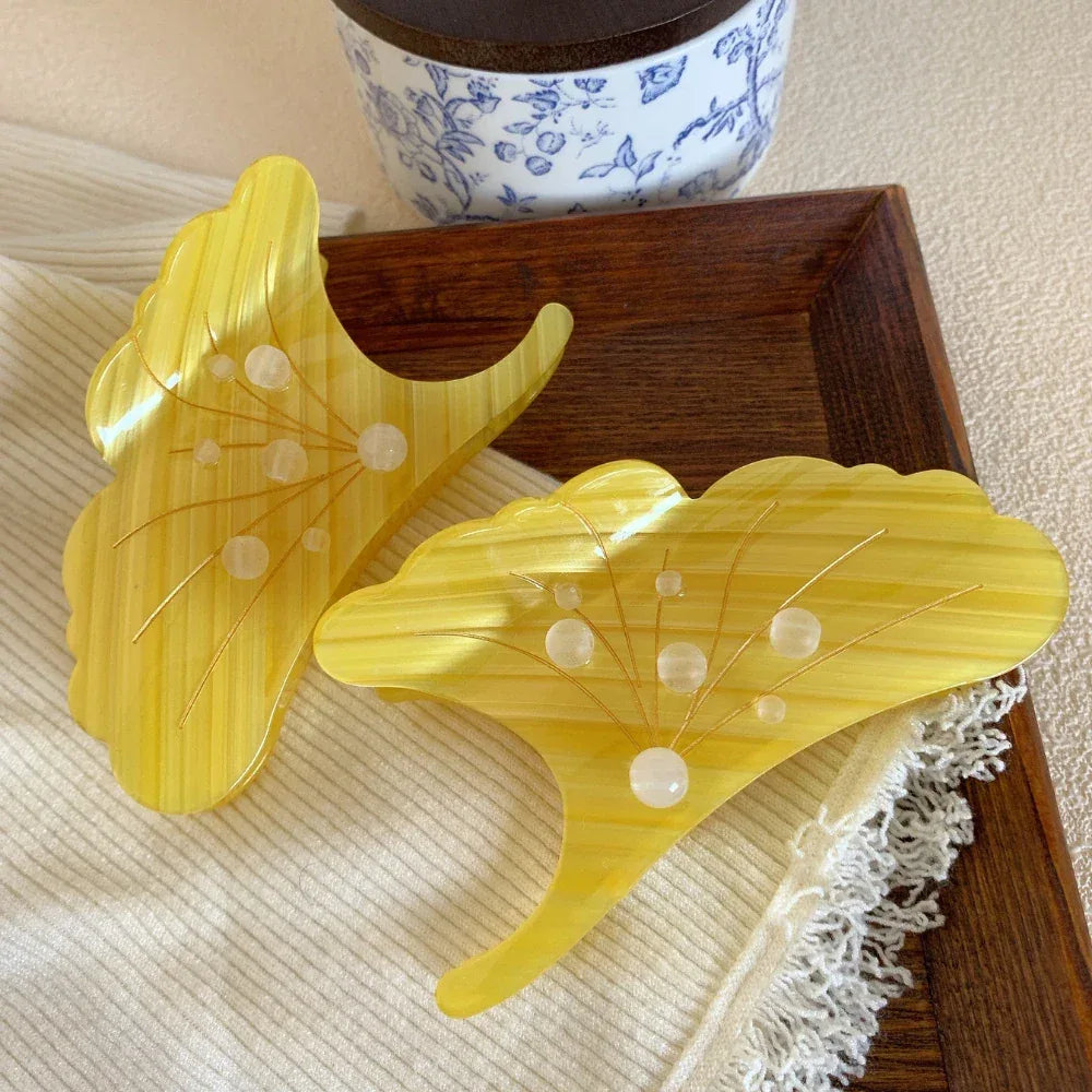 Two yellow ginkgo leaf-shaped hair claw clips arranged on a textured white cloth with lace trim, with a decorative blue and white ceramic pot in the background.