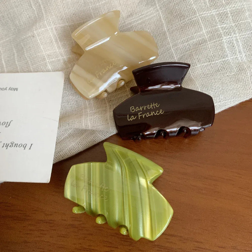Three acetate claw hair clips in beige, green, and brown on a textured cloth.