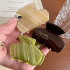 A hand holding three acetate claw hair clips in beige, green, and brown.