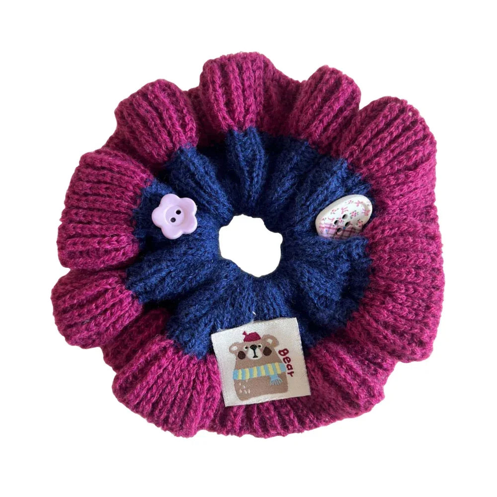 Close-up of a pink and blue knitted flower scrunchie with a bear tag and button embellishments.