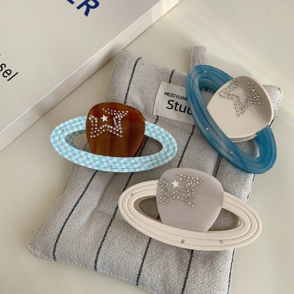 Three planet-shaped hair clips with rings and star embellishments, displayed on a grey fabric.