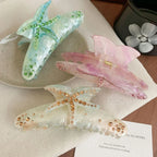 Three decorative hair claw clips arranged on a textured surface. One is blue with green rhinestones, one is pink with pink rhinestones, and one is white with gold rhinestones. All feature a starfish design.