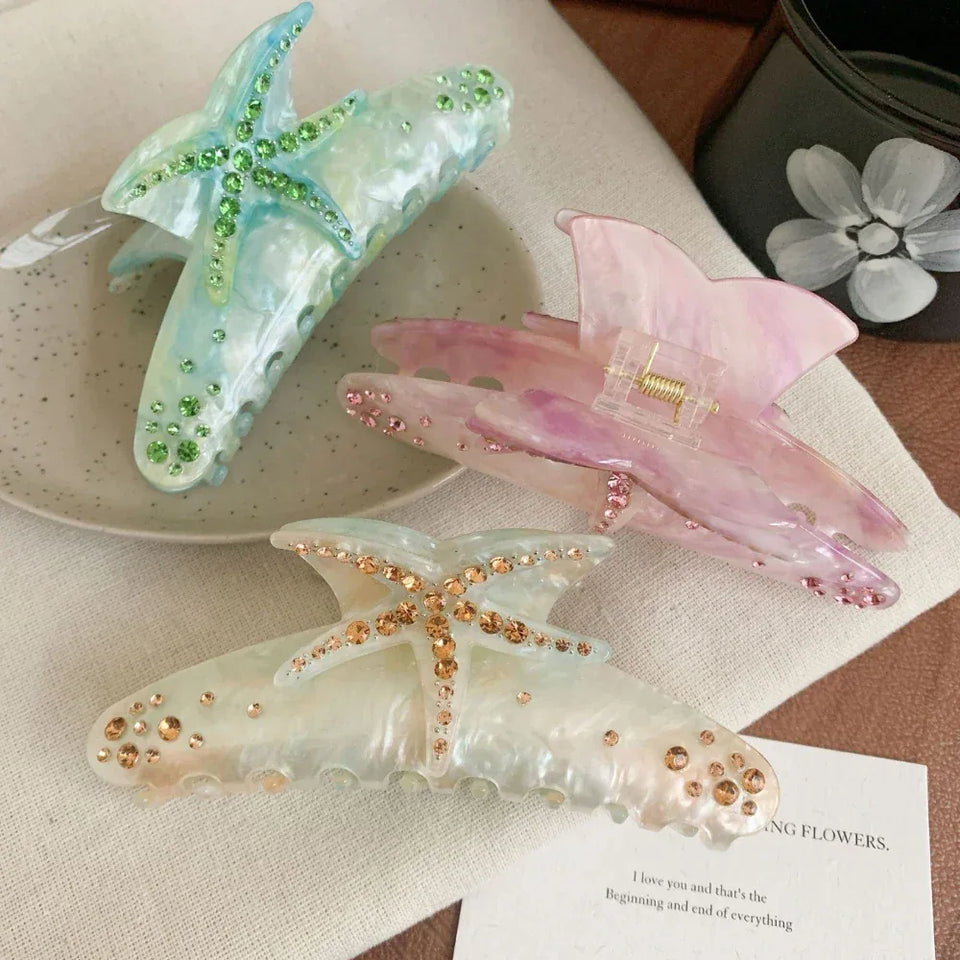 Three decorative hair claw clips arranged on a textured surface. One is blue with green rhinestones, one is pink with pink rhinestones, and one is white with gold rhinestones. All feature a starfish design. Secondary image
