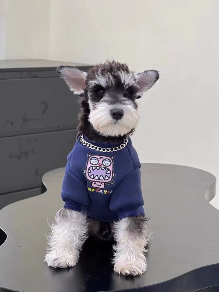 Mini Schnauzer puppy wearing a navy blue sweatshirt with a pink monster graphic on the front and a chain detail around its neck, sitting on a black table.