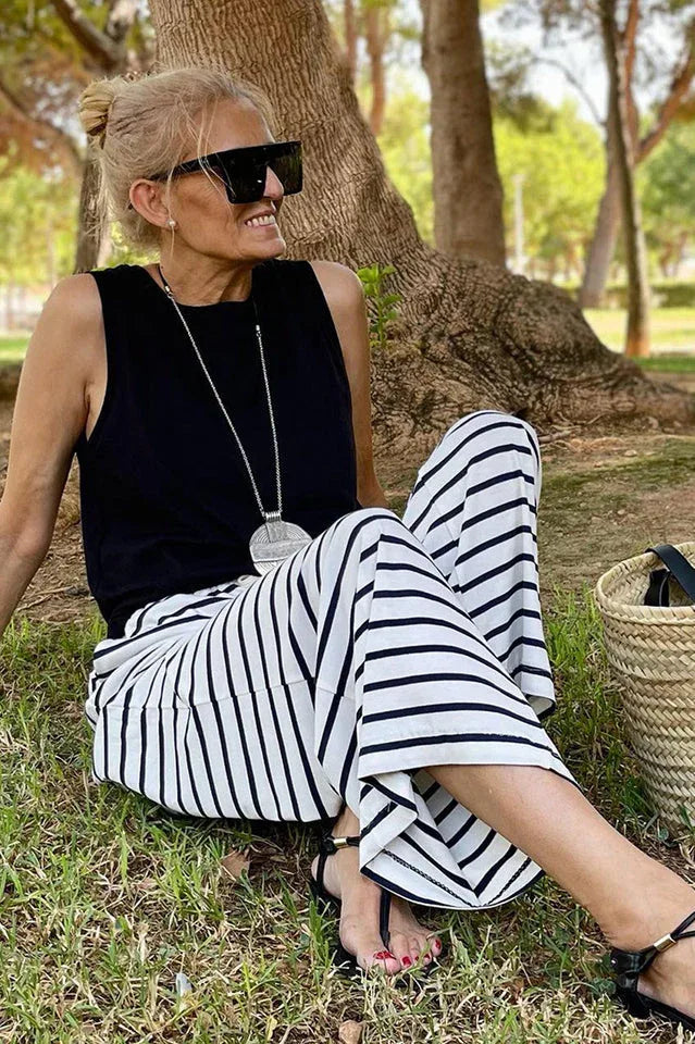 Woman sitting on the grass wearing a black sleeveless top and black and white striped wide leg pants, with a straw tote bag beside her.