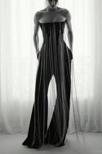 Model wearing a black strapless jumpsuit with long fringe detailing and wide legs, standing in front of a window.
