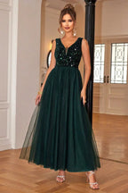 Woman in a floor-length dark green sequined and tulle formal dress with a v-neckline, posing with hands on hips.