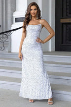 Woman wearing a floor-length, spaghetti strap, sequined white dress with a high slit, standing on stairs.