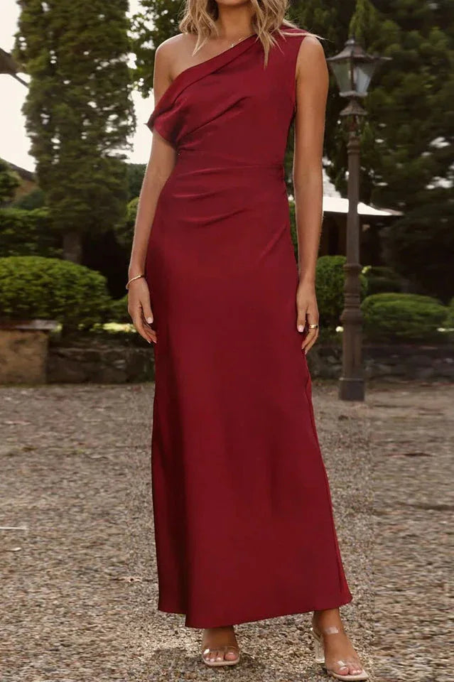 Woman standing in a garden wearing a burgundy one-shoulder satin maxi dress.