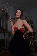 Woman holding a drink, wearing a black velvet strapless mini dress with red heart bust details, in front of a Christmas tree.