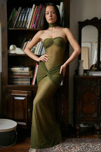 Woman wearing a strapless, shimmering olive green gown with ruched detailing and a knotted front accent, standing in front of a bookshelf.
