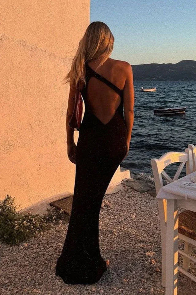 Woman from behind wearing a black sleeveless floor-length gown with a unique strap detail across the back, standing by the water at sunset.
