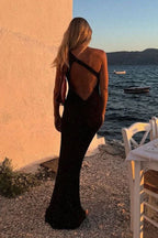 Woman from behind wearing a black sleeveless floor-length gown with a unique strap detail across the back, standing by the water at sunset.