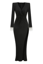 Full-length studio shot of a long-sleeved black jersey knit gown with a plunging V-neckline, ruched bodice, thigh-high slit, and embellished cuffs.