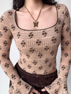 Close-up of a fitted, long-sleeved square neck top with an allover brown floral print on a beige textured knit fabric. The neckline and sleeve cuffs have dark scalloped trim. A butterfly pendant necklace is worn.