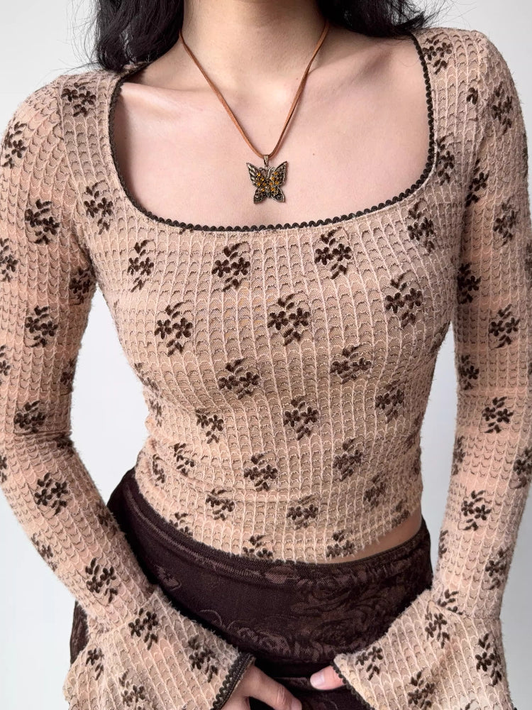 Close-up of a fitted, long-sleeved square neck top with an allover brown floral print on a beige textured knit fabric. The neckline and sleeve cuffs have dark scalloped trim. A butterfly pendant necklace is worn.