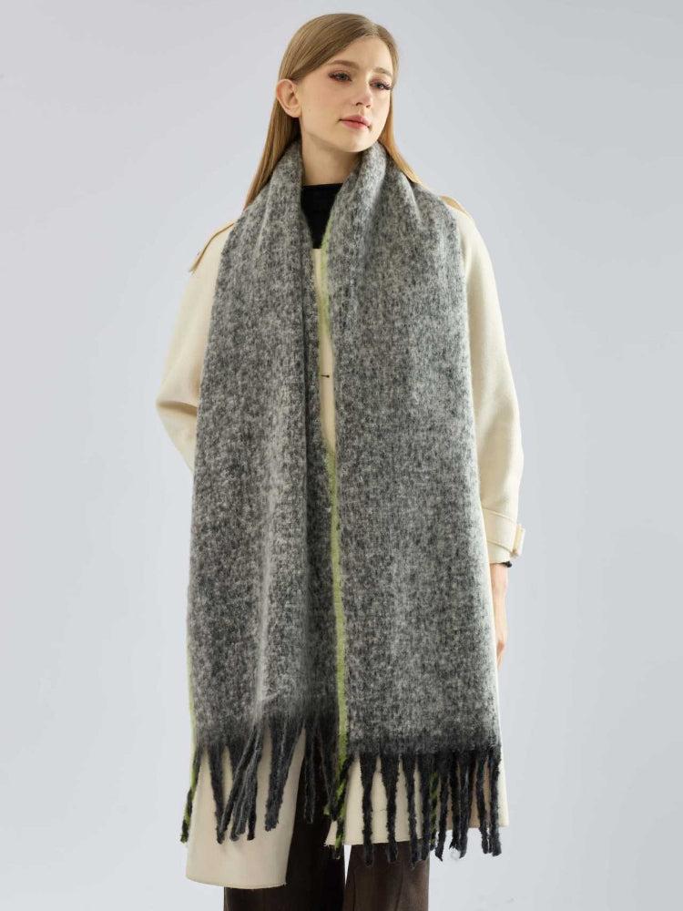 Full body shot of a woman in a cream coat and a long, grey, textured knit scarf with fringe and lime green trim.