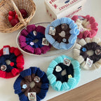 An overhead shot of several knitted flower scrunchies, some on a tray and some on a table, with a basket of apples in the background.