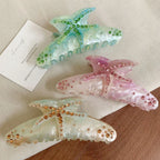 A top-down view of three stylish hair claw clips with starfish motifs and rhinestone accents, set against a light-colored fabric and a small card with text.