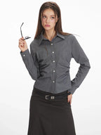 Woman wearing a grey long-sleeved button-down shirt with ruching detail, holding her glasses.
