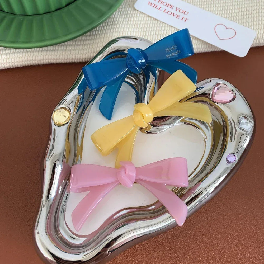 A heart-shaped silver dish holding three resin bow-shaped hair clips in blue, yellow, and pink.